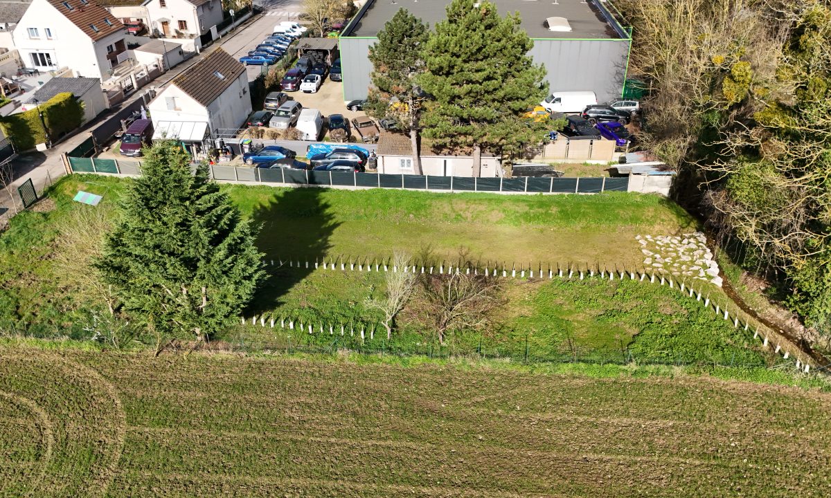 Un petit ruisseau vu du ciel longeant un champ, une forêt et des maisons